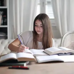 Dziewczynka z długimi brązowymi włosami pisze w otwartych książkach przy drewnianym stole.