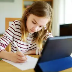 Dziewczynka w bluzie w paski pisze coś na kartce, obok tablet i kredki.