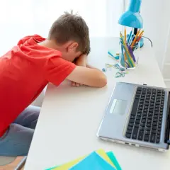 Chłopiec śpi na biurku obok laptopa i przyborów szkolnych.
