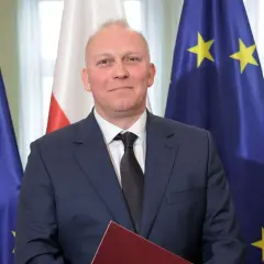 Tomasz Szabłowski w garniturze, na tle flag Polski i Unii Europejskiej.