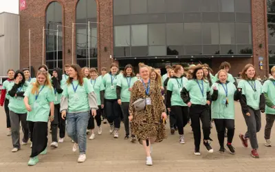 Grupa młodych ludzi w zielonych koszulkach idzie przed budynkiem z cegły.