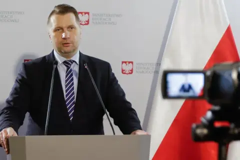 Mężczyzna w garniturze przemawia przy mównicy na tle flagi Polski i logo Ministerstwa Edukacji Narodowej.