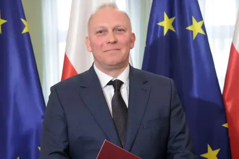 Tomasz Szabłowski w garniturze, na tle flag Polski i Unii Europejskiej.