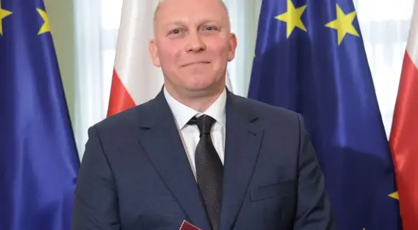 Tomasz Szabłowski w garniturze, na tle flag Polski i Unii Europejskiej.