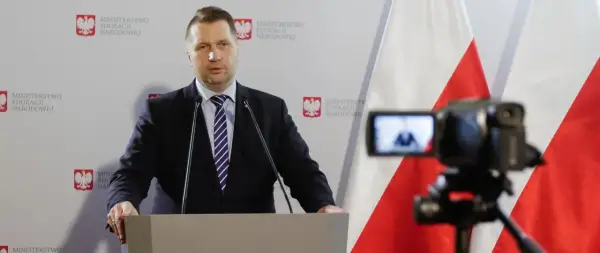 Mężczyzna w garniturze przemawia przy mównicy na tle flagi Polski i logo Ministerstwa Edukacji Narodowej.
