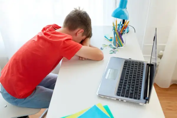 Chłopiec śpi na biurku obok laptopa i przyborów szkolnych.